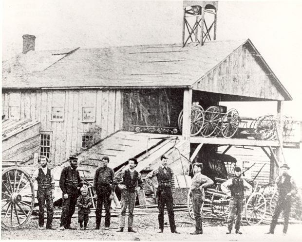 Image en noir et blanc représentant huit hommes et un jeune garçon alignés devant un bâtiment en bois, entourés de chariots et de pièces pour ceux-ci.