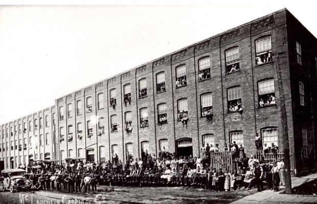 Plusieurs centaines d’ouvriers posent avec quatre automobiles devant une usine imposante.
