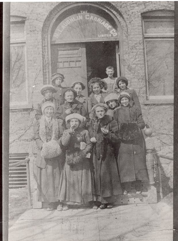Des employées de bureau portant des vêtements typiques des années 1900 se tiennent sur les marches d’un bâtiment en brique sur lequel on peut lire l’inscription « The McLaughlin Carriage Co Limited » peinte au-dessus de la porte.