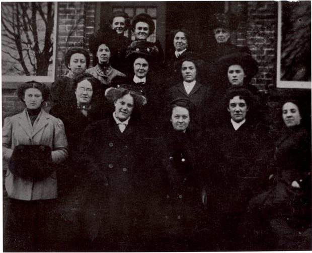 Quinze employées de bureau en tenue formelle des années 1900 se tiennent sur les marches d’un bâtiment en brique.