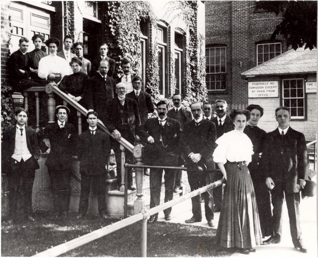 Plusieurs dizaines d’employés et employées de bureau en tenue formelle des années 1900 se tiennent sur les marches d’un bâtiment recouvert de lierre.