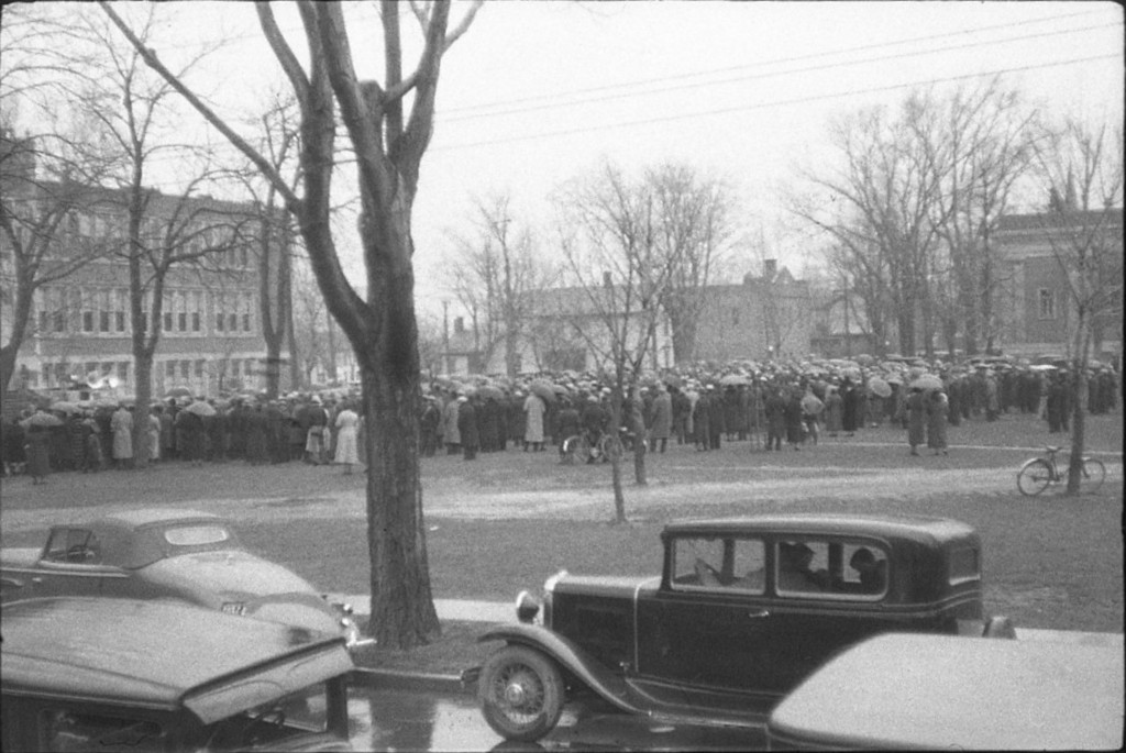 Image en noir et blanc représentant plusieurs centaines de personnes avec des parapluies rassemblées dans un parc, avec de vieilles voitures garées au premier plan.