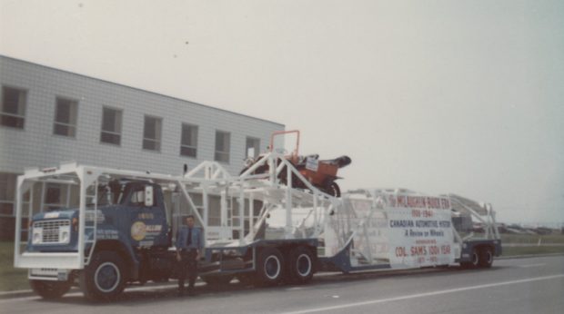 Image en couleur d’une voiture ancienne sur le plateau d’un camion de transport devant un bâtiment industriel. Un homme en uniforme d’agent de sécurité se tient devant le camion.