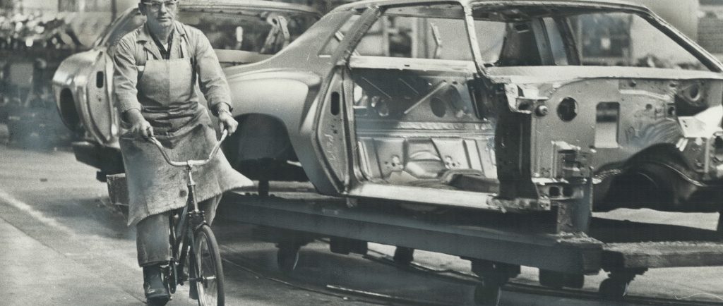 Image en noir et blanc représentant un homme portant un tablier et une casquette de baseball à l’envers roulant à vélo à côté d’une voiture inachevée sur une chaîne de montage.