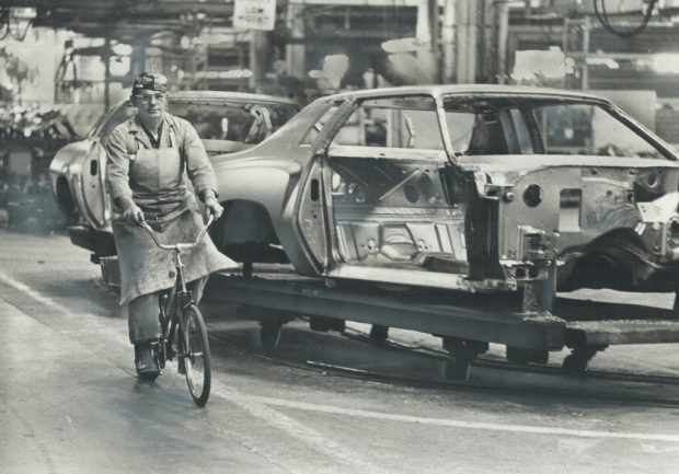 Image en noir et blanc représentant un homme portant un tablier et une casquette de baseball à l’envers roulant à vélo à côté d’une voiture inachevée sur une chaîne de montage.