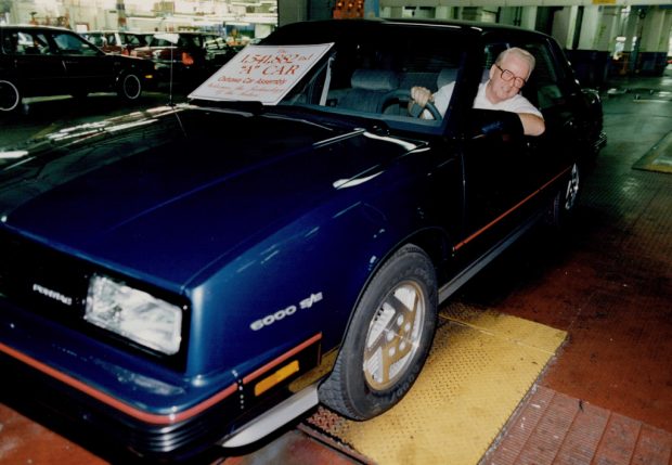 Photo en couleur d’un homme conduisant une voiture le long de la chaîne de montage d’une usine. Un écriteau sur le pare-brise indique « La 1 541 882e voiture A de la chaîne de montage d’Oshawa ».