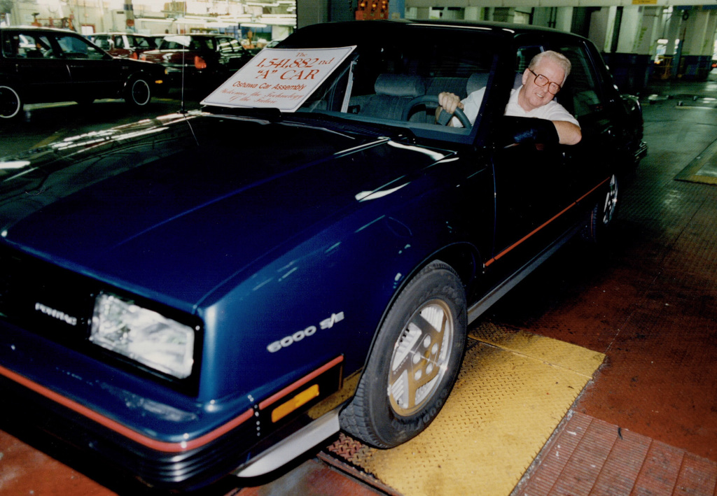 Photo en couleur d’un homme conduisant une voiture le long de la chaîne de montage d’une usine. Un écriteau sur le pare-brise indique « La 1 541 882e voiture "A" de la chaîne de montage d’Oshawa ».