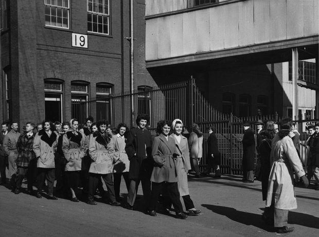 Photographie noir et blanc de plusieurs dizaines de femmes et d’hommes défilant devant une usine.