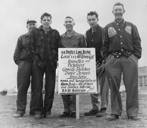Image en noir et blanc représentant cinq hommes debout autour d’une pancarte présentant une revue sur le piquet de grève de 55.