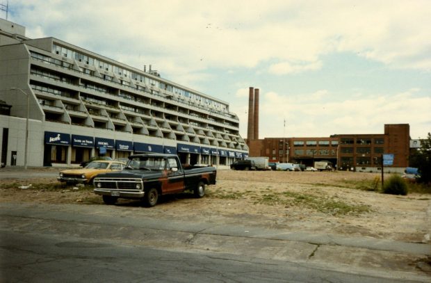 Image en couleur d’un immeuble résidentiel en construction à côté d’une usine. Plusieurs voitures et camions des années 70 sont garés devant l’appartement.