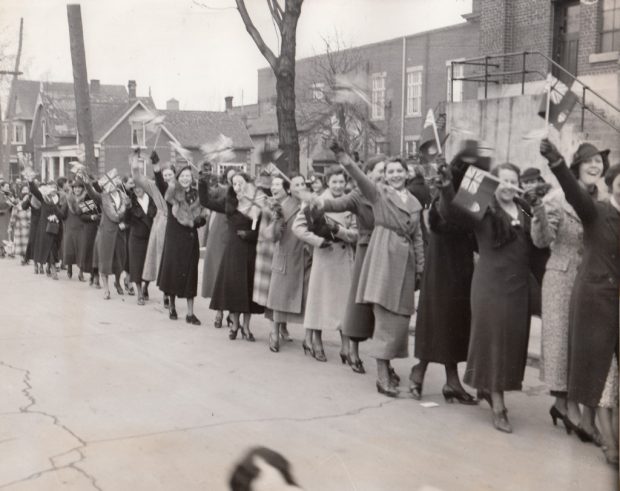 Un groupe de femmes défile devant un bâtiment industriel, brandissant des drapeaux canadiens et britanniques.