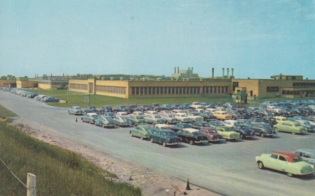 Sur cette photographie, plusieurs centaines de voitures anciennes sont garées devant une usine.
