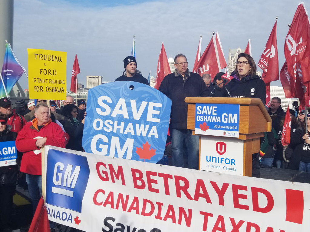 Une femme donne un discours depuis un podium sur lequel il est marqué « SAUVEZ GM OSHAWA », devant plusieurs dizaines de manifestants portant des drapeaux. Un panneau écrit à la main en arrière-plan indique « TUREAD & FORD COMMENCENT À SE BATTRE POUR OSHAWA ».