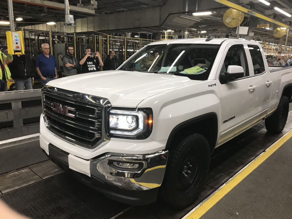 Un pick-up traverse une zone industrielle. Plusieurs hommes et femmes se tiennent à l’arrière-plan et prennent des photos avec leurs téléphones portables.