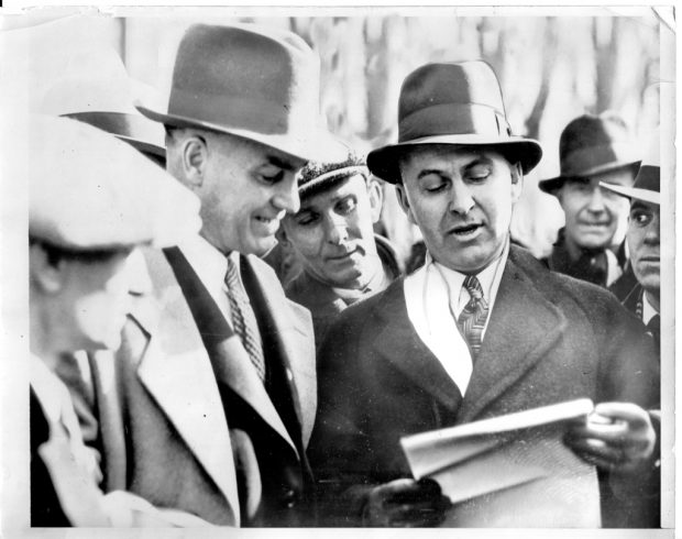 Plusieurs hommes en habits de 1930 discutent du contenu d’une feuille de papier.
