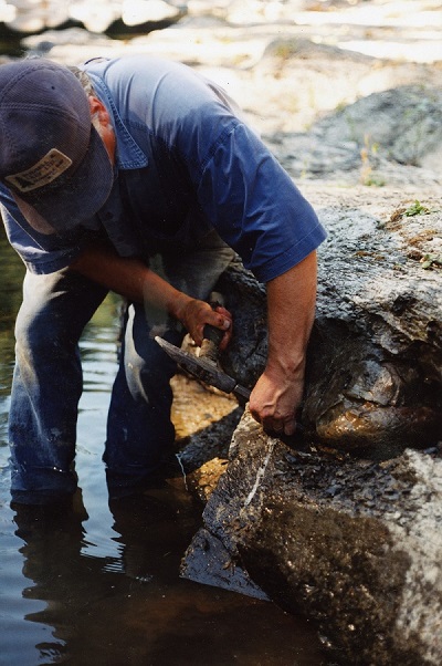 Graham Beard, penché au-dessus d’un rocher au milieu de la rivière, marteau et burin à la main.