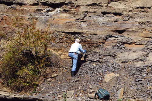 Graham Beard, debout au bord d’une rivière où des couches de roches sédimentaires sont visibles.