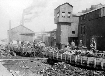 Les locomotives à vapeur transportaient des wagons remplis de matières premières. Amas de déchets en avant plan et ouvriers se tiennent de bout derrière des wagons.