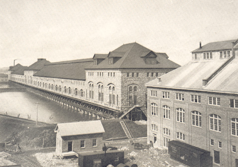 La centrale en pierre de grès et deux autres bâtiments plus petits en avant plan. On aperçoit la rivière St. Mary à la droite de la centrale.