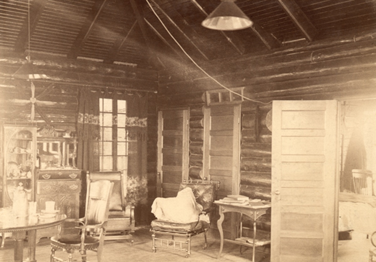 Intérieur du blockhaus, fait en bois rond. On y voit une fenêtre, une lampe suspendue au plafond, une table, plusieurs chaises et une commode.