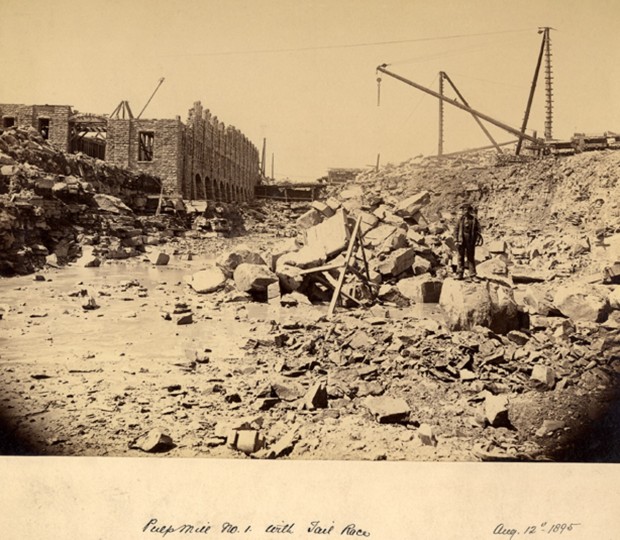 Du gravier et des roches sont éparpillés partout sur le chantier. En arrière plan, on distingue des grues, un ouvrier et un édifice en construction.