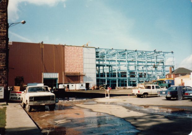 Structure d'acier érigée sur un des côtés du nouvel édifice ; l'autre façade à gauche est recouverte d'un parement. Des camions sont présents et un écriteau appuyé sur l'atelier d'usinage indique «chantier de construction».