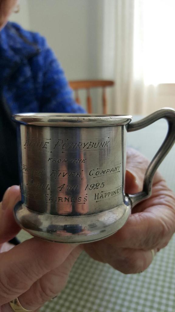 Deux mains de vieille femme qui tiennent une petite tasse en argent portant l'inscription : «To Annie Pohrybunk, April 4, 1925 from the Spanish River Company».