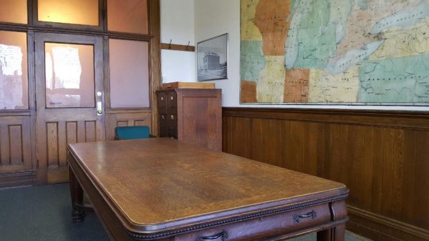 Grande table de travail en bois installée au centre d'une salle lambrissée, positionnée en face de la porte d'entrée. Grande murale illustrant une carte géographique sur la droite.