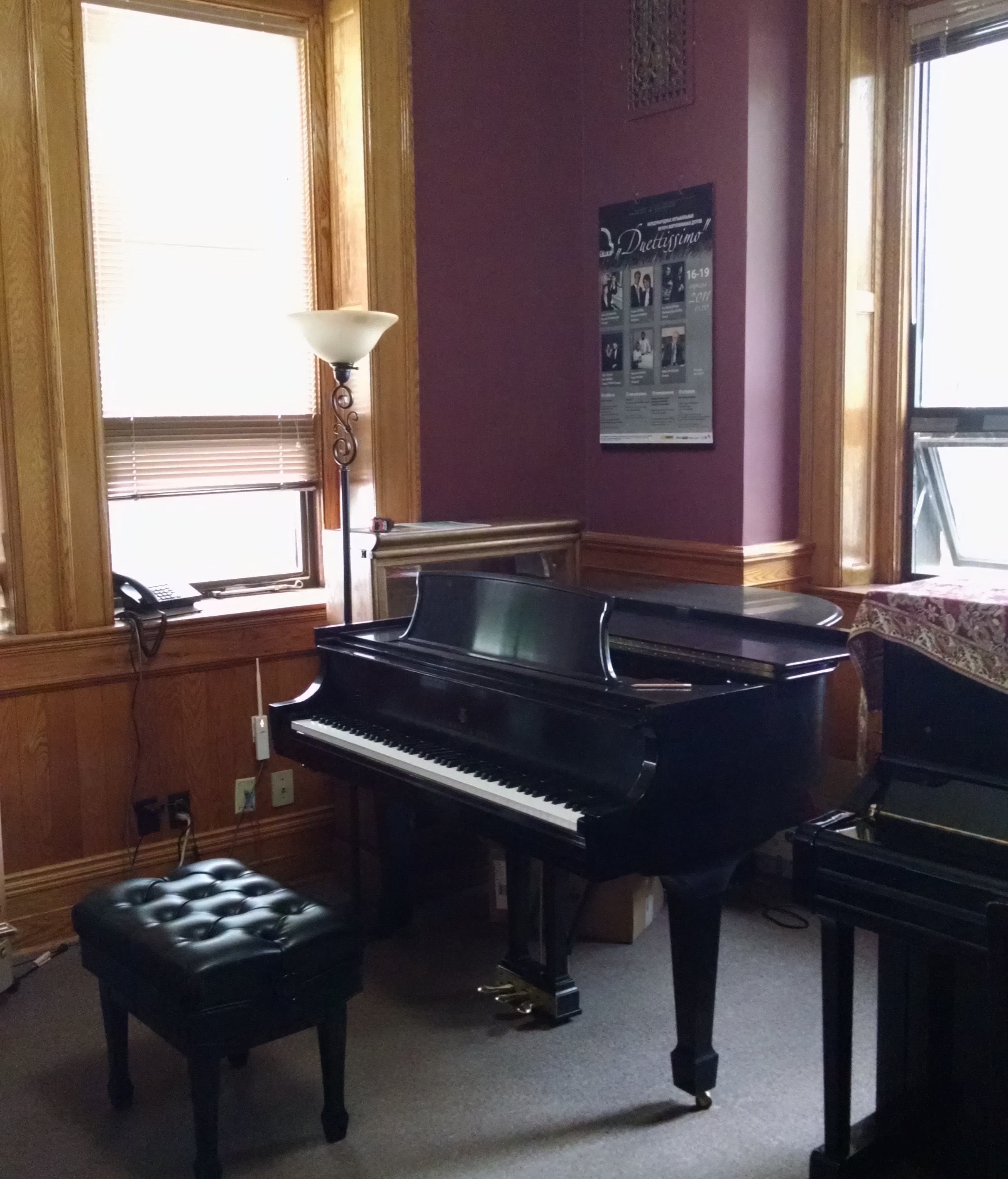 Deux pianos et un banc installés côte-à-côte dans une petite salle peinte de couleur bourgogne, avec de grandes fenêtres ; lambrissé, porte et plinthe en chêne ; tapis mur à mur.