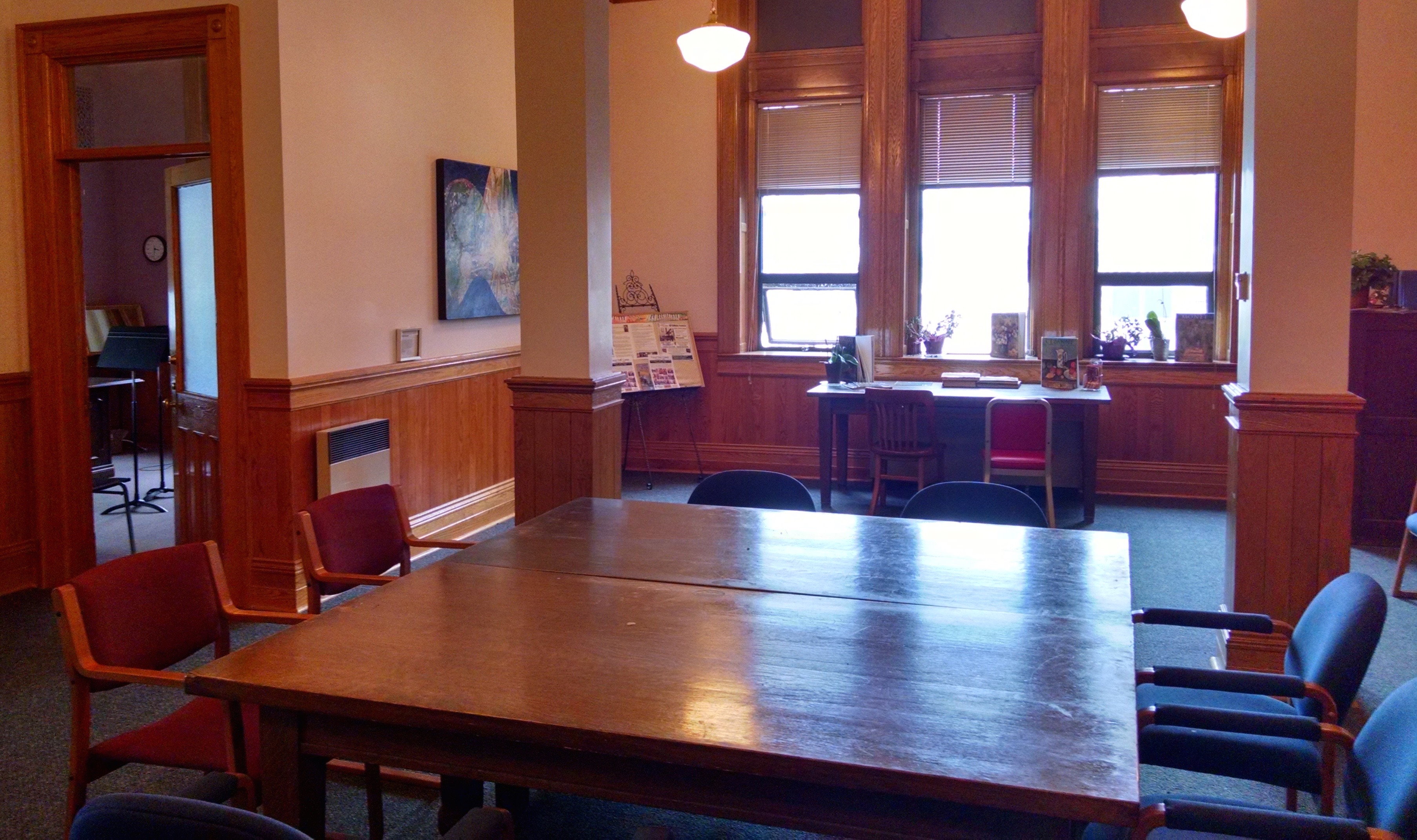 Vue en direction ouest dans la salle d'attente du Conservatoire de musique d'Algoma. Photo prise de la porte d'entrée. Grosses poutres de support au centre de la salle. Large fenêtre directement en face. Porte ouverte à gauche donnant sur un studio ou un bureau. Grande table et chaises au centre de la salle.