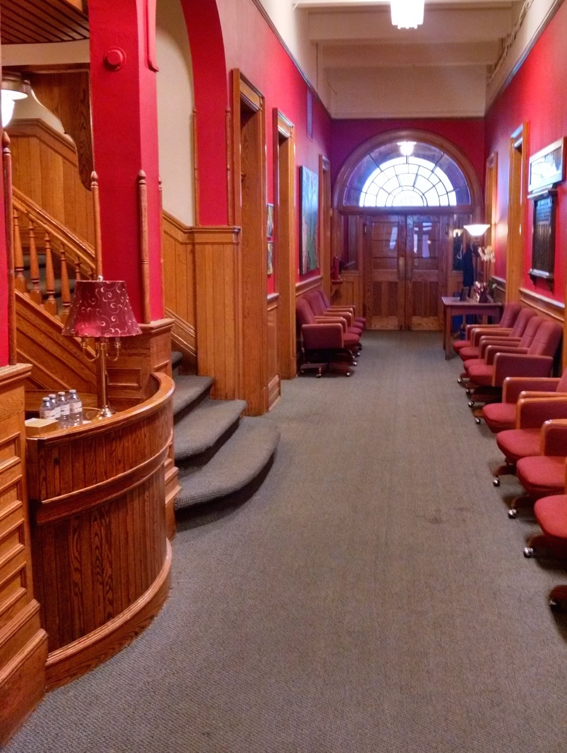 Hall d'entrée et escalier central du Conservatoire. Rangées de chaises alignées de chaque côté du hall d'entrée. Portes françaises et fenêtres au fond du hall.  Détails de l'escalier et du bureau de réception en bois.