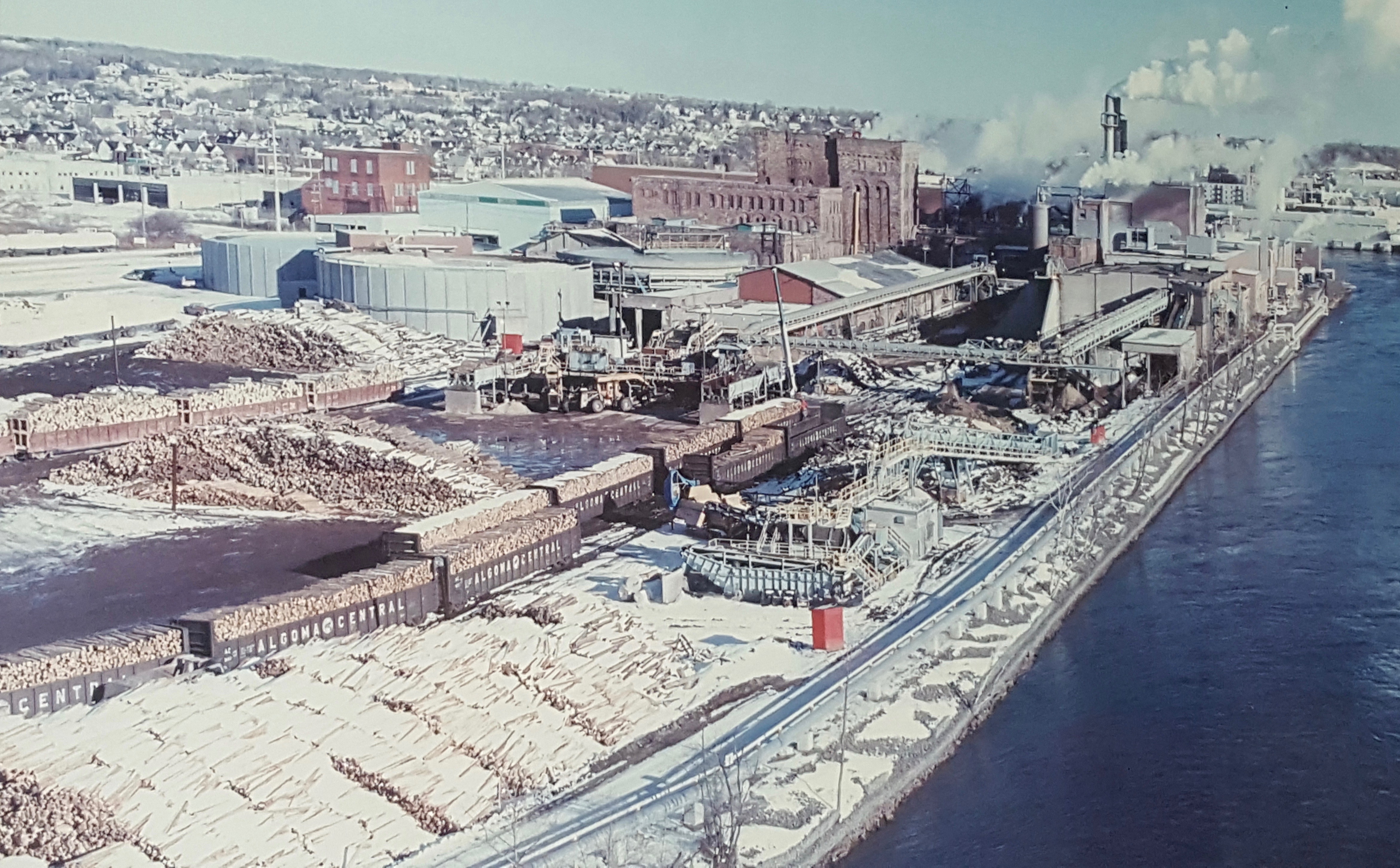 Photo d'une photographie encadrée. Vue du haut du site de la papetière avec édifices, amoncellements de billots de bois et wagons de train.