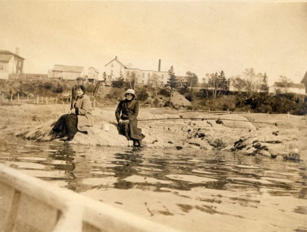 Photo sépia (vers 1910) : deux femmes assises sur un rocher au bord de l’eau. Arrière-plan : rive (herbe, arbres, bâtiments). Premier plan : une partie d’embarcation (photo prise à bord d’un bateau). Scène côtière/riveraine.