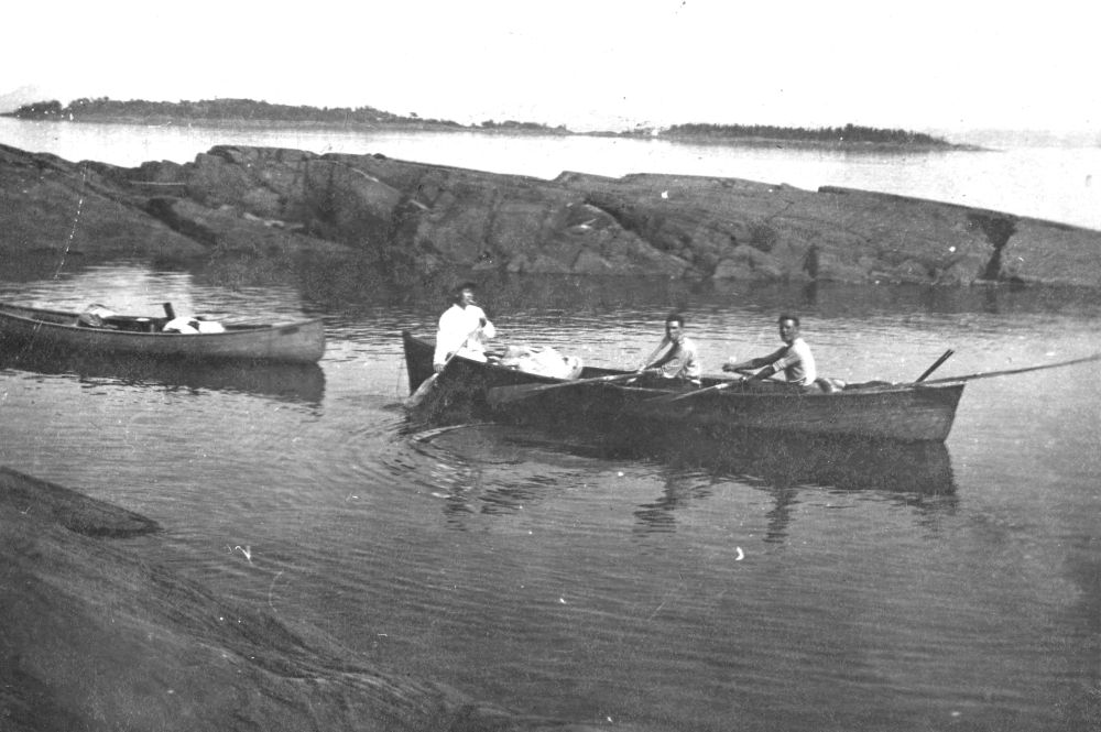 Photo en noir et blanc (années 1910) : trois personnes dans une embarcation en eau calme près d’une côte rocheuse. Premier plan à droite : canot de bois (trois personnes rament, vêtements clairs). Gauche : autre canot. Arrière-plan : côte rocheuse accidentée, îles/terres lointaines horizon. Scène à Kamouraska.