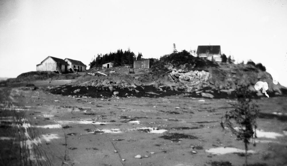 Photo en noir et blanc (vers 1920) : paysage de l’Île aux Patins, Kamouraska. Terrain accidenté (rochers, végétation clairsemée, conifères). Plusieurs petits bâtiments en bois dispersés. Premier plan : zone plus plate (grève/partie basse, traces d’eau/marée basse).