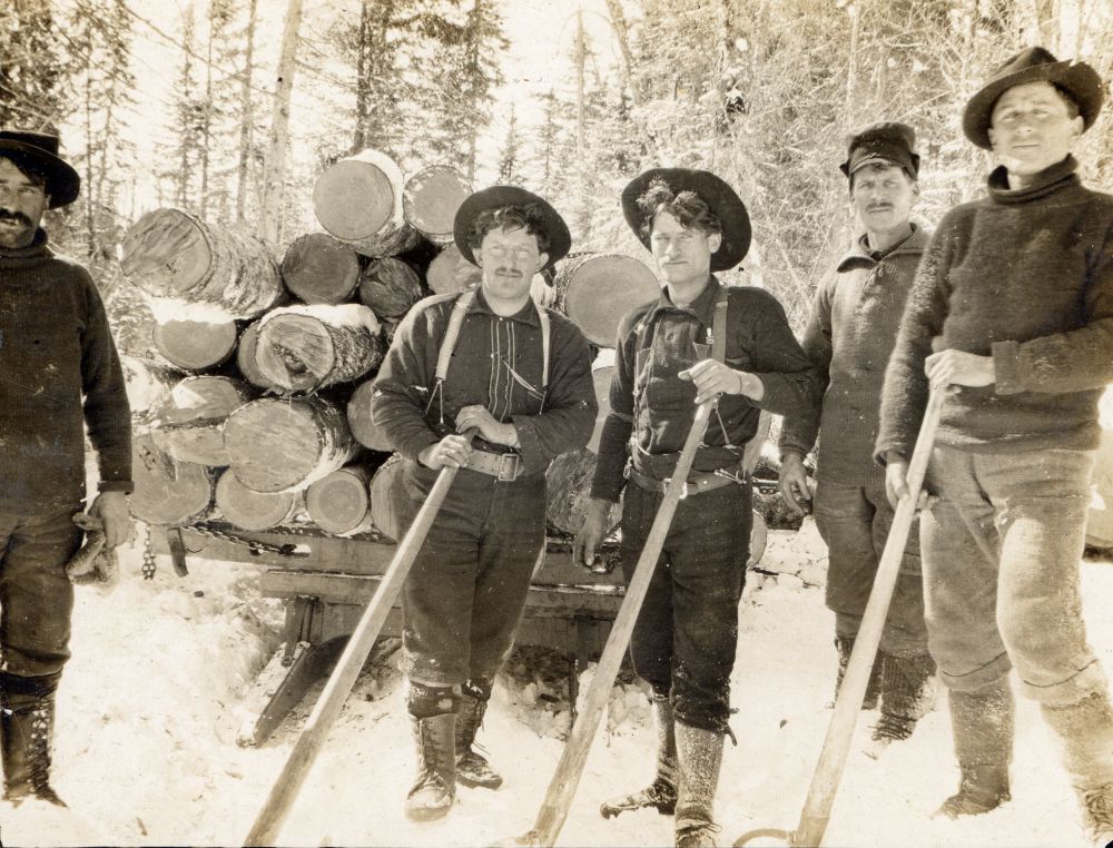 Photo sépia d’un chantier forestier : cinq hommes posent dans environnement enneigé. Ambiance de dur labeur hivernal. Vêtements chauds, bottes hautes. Quatre des hommes ont des chapeaux, chacun tenant une gaffe de drave. Derrière : grande pile de billes de bois sur un traîneau. 