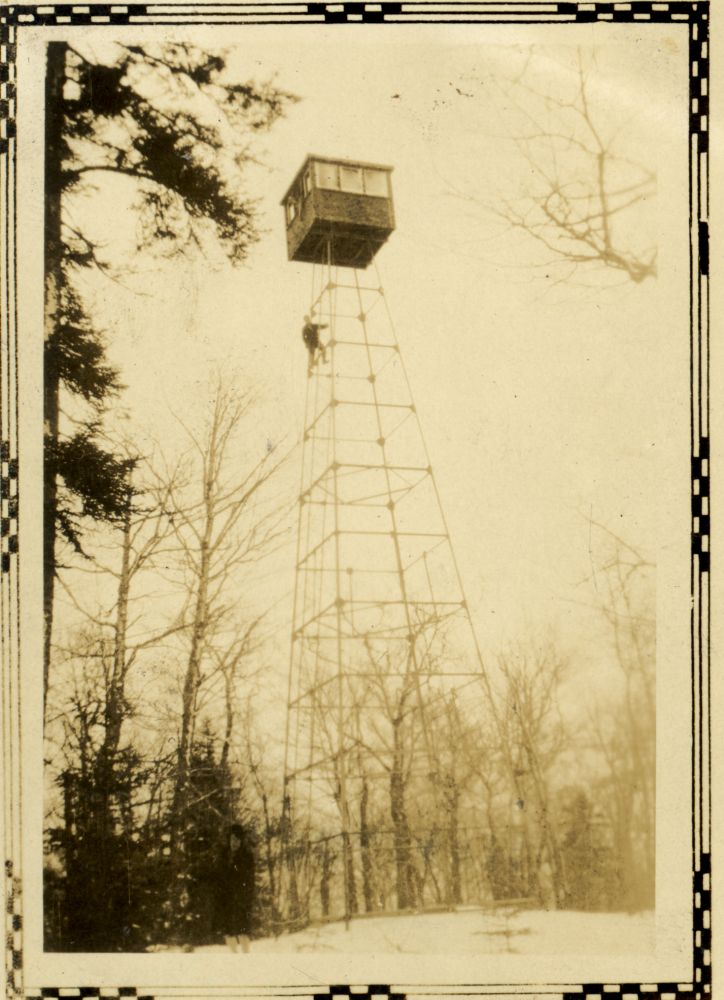 Photo sépia : haute tour à feu en métal, forêt hivernale, arbres dénudés.  Au sommet de la tour : petite cabine carrée avec fenêtres, une personne grimpant sur la structure est visible. Sol enneigé, quelques conifères. Ciel uniforme et clair. Bordure décorative.