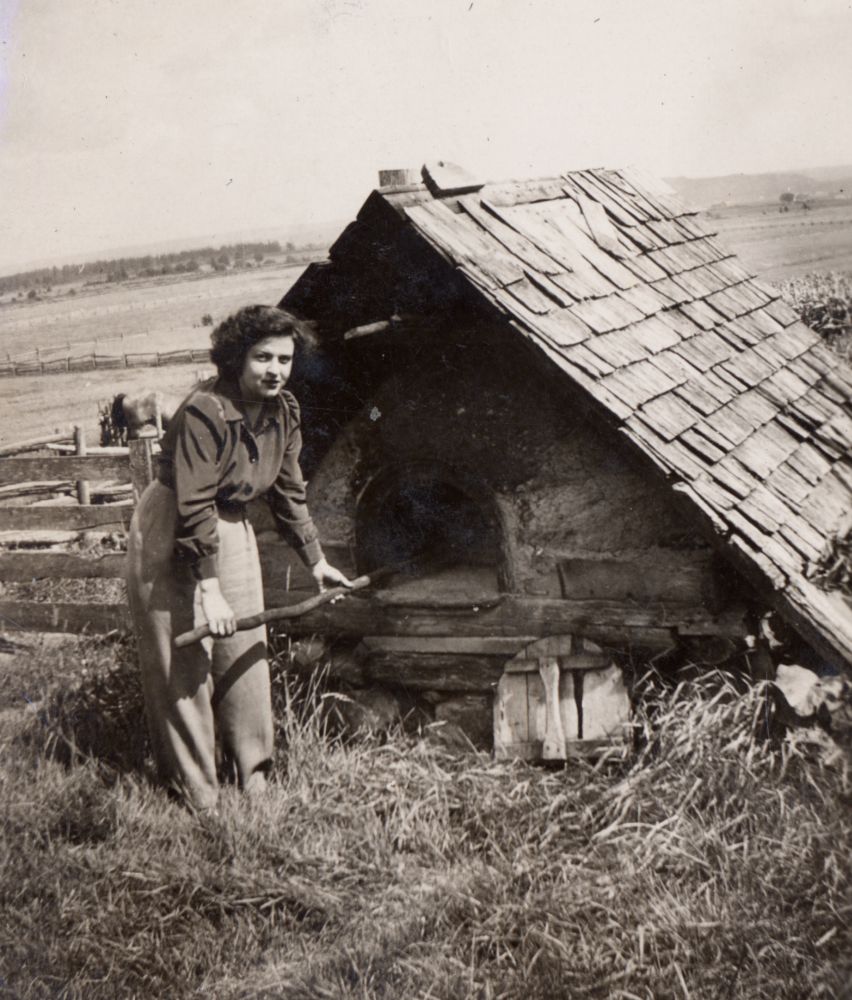Photo en noir et blanc : femme debout près d’un four à pain extérieur. La femme tient un long bâton en bois. Le four en bois et en pierres, possède un toit de bardeaux usés. Clôture en arrière-plan, paysage rural lointain. Ambiance d’époque de cuisson extérieure. 