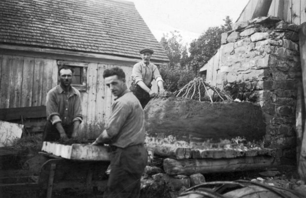 Photo en noir et blanc (années 1930) : trois hommes construisent un four à pain extérieur constitué de branches recouvertes de terre ou de mortier. Derrière : bâtiment en bois avec un toit en bardeaux. Scène de construction artisanale d’un four à pain extérieur.