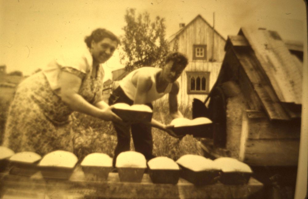 Photo sépia : une femme et un homme sortent des moules à pain du four extérieur. Devant: four et autres moules à pain remplis. Arrière-plan : maison en bois et végétation