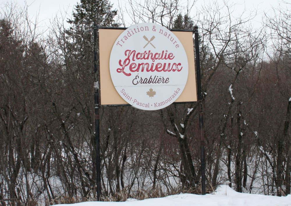 Panneau beige avec une bordure brune et un cercle blanc central. Inscription "Tradition & nature" en arc au-dessus. Nom rouge "Nathalie Lemieux" au-dessus "Érablière". Bas : "Saint-Pascal • Kamouraska" et feuille d’érable. Environnement hivernal enneigé, arbres dénudés.