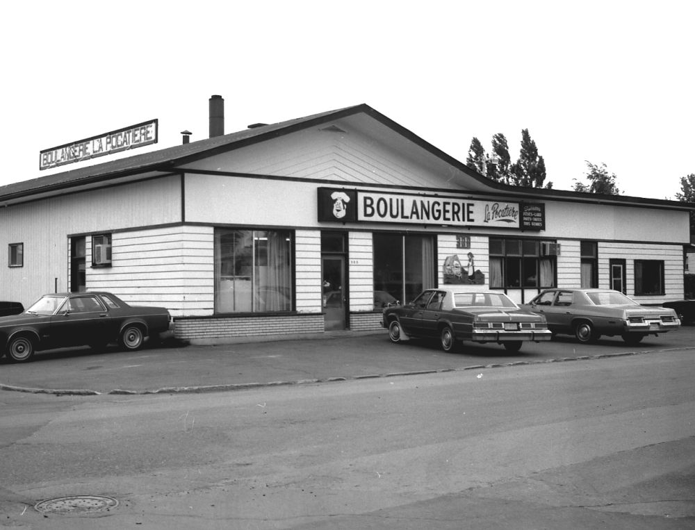 Photo en noir et blanc : façade d’un bâtiment commercial "BOULANGERIE ". Bâtiment plain-pied, revêtement clair horizontal, toit plat pente légère. Grandes vitrines. Plusieurs voitures stationnées devant (pavé). Photo début des années 1970.