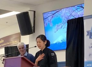 Asian woman with black hair, black chef's uniform, speaking into a microphone, hand on chest. To the left, an older man with gray hair. Behind: large screen with cherry blossoms. Atmosphere of a speech at an event.