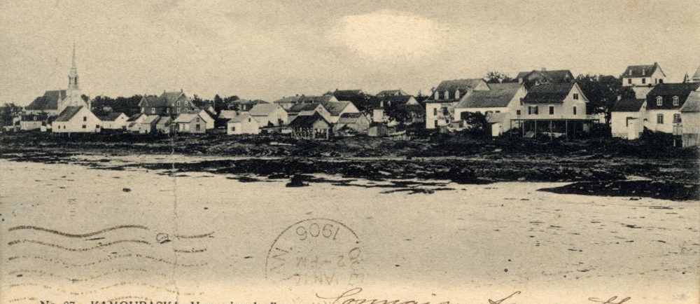 Carte postale sépia No. 87. KAMOURASKA. – Vue prise du fleuve (panoramique d’une petite municipalité côtière). Premier plan : étendue d’eau (fleuve Saint-Laurent). Sur la rive : maisons, église au clocher pointu. Inscriptions manuscrites en français. Timbre 1906. En bas à gauche : Editeurs: Pruneau & Kirouac, Québec..