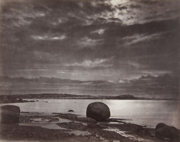 Épreuve albumine (1876, Alexander Henderson) : Le soir sur la plage à Métis (paysage marin mélancolique crépuscule). Ciel dominant (nuages sombres texturés, trouées de lumière). Premier plan : plage rocailleuse humide (gros rochers sombres dont un bloc arrondi au centre droit). 