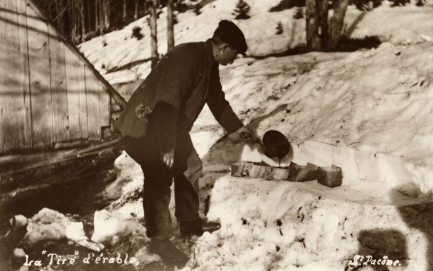 Photo sépia la Tire d'érable (S. Pacôme) : homme en habits d’hiver versant de la tire d'érable dans des récipients d’écorce sur la neige. Arrière-plan : boisé enneigé, une partie d’une structure en bois à gauche.