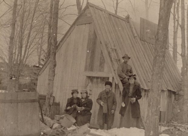 Photo sépia : célébration familiale devant une cabane en bois rustique (environnement boisé et enneigé). Premier plan gauche : deux jeunes femmes assises. Centre : jeune homme debout. Droite : femme debout. Un homme assis sur le toit de la cabane. Extrême gauche : grand tonneau en bois.