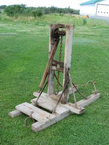 Stake puller on a lawn. Vertical wooden structure, metal chain, long side arm, front platform with hook. Background: vegetation, white farm building with green roof.