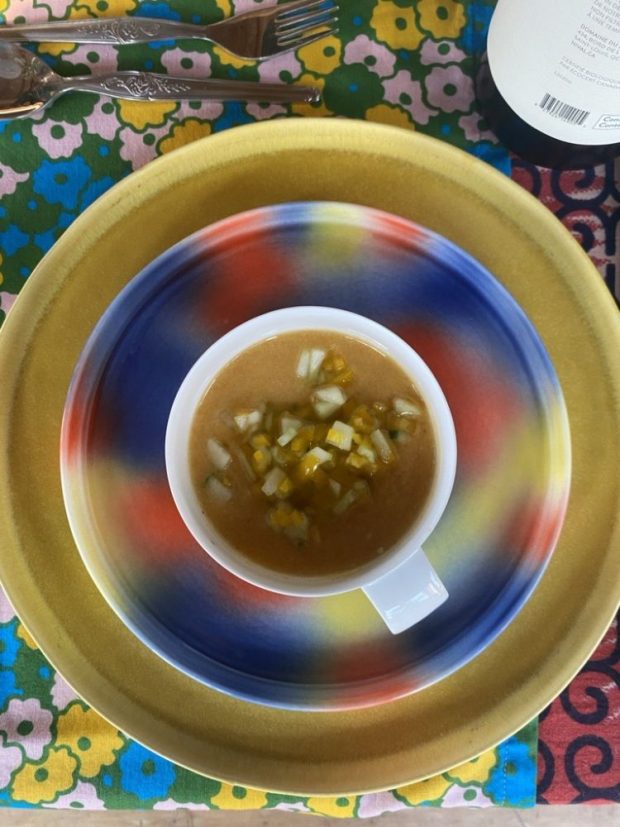 Plan de dessus sur un bol de soupe orangée posé sur une petite assiette à motifs bleus, rouges et jaunes, elle-même placée sur une grande assiette dorée. L'ensemble est sur une nappe à motifs floraux colorés.