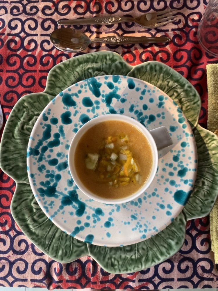 Plan de dessus sur un bol de soupe orangée garnie de petits dés de légumes, posé sur une assiette décorative blanche mouchetée de taches bleues, elle-même placée sur une grande assiette en forme de feuille verte. L'ensemble repose sur une nappe rouge et bleue à motifs.